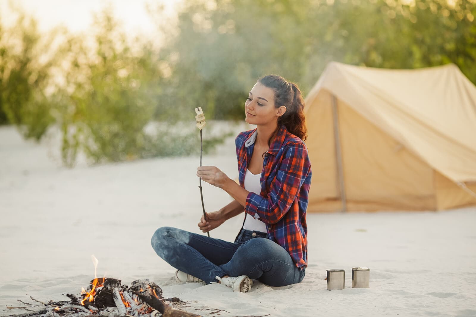 Young woman solo camping cooking snacks on campfire, on beach in front of cream coloured tent